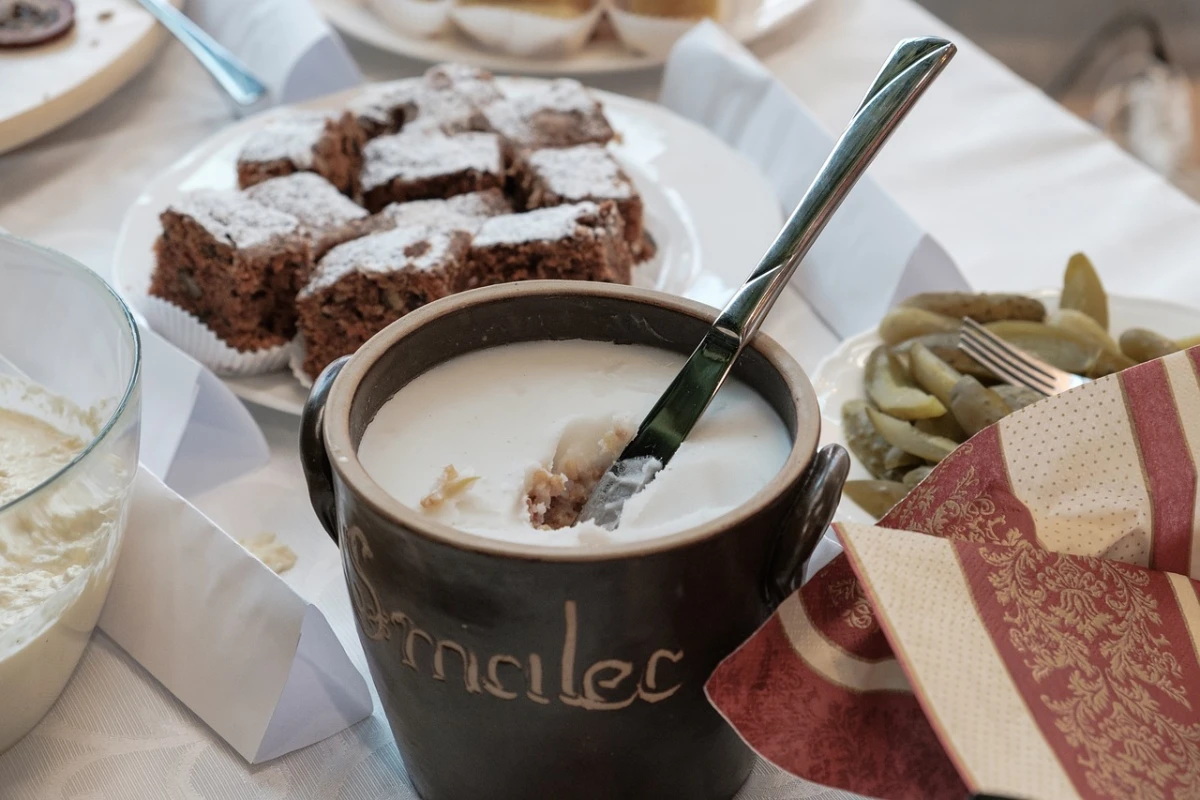 Gliniany garnek z gęsim smalcem i łyżką, stojący na stole wśród innych potraw, w tle talerz z ciastem i miska z ogórkami